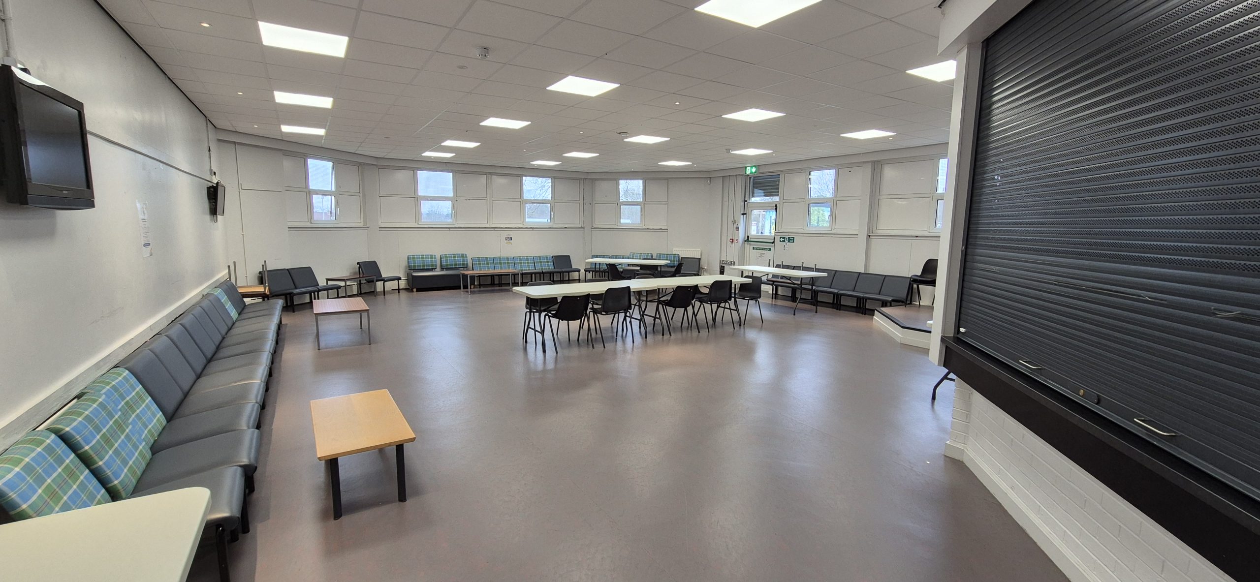 Coffee Bar at Whitburn Community Centre — a bright, sociable lounge space with perimeter bench seating upholstered in tartan fabric, an open floor area, and wall-mounted TV screens